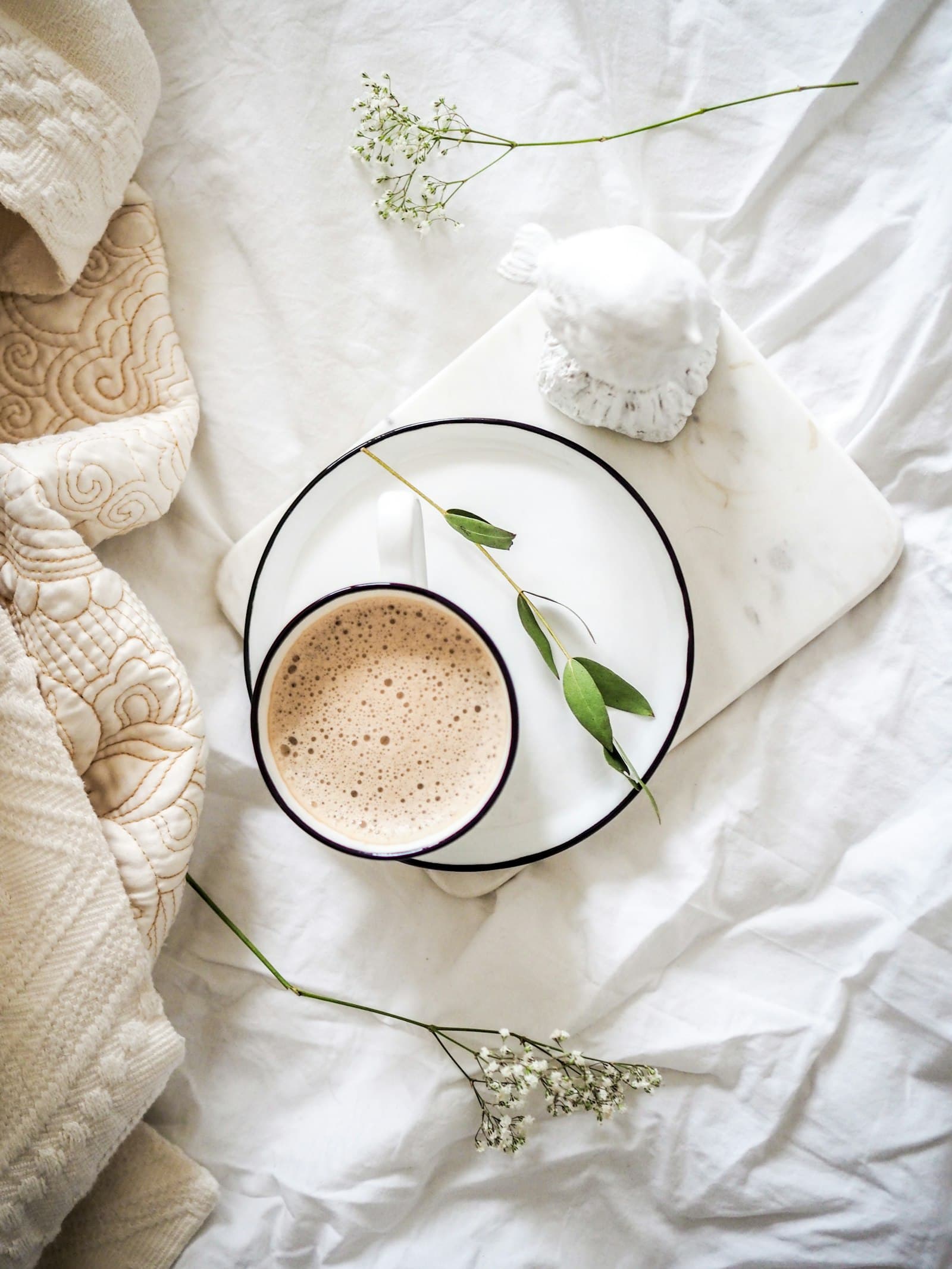 Tasse de café sur un plateau de marbre posé sur du linge blanc, avec un plaid crème et des brins de gypsophile.