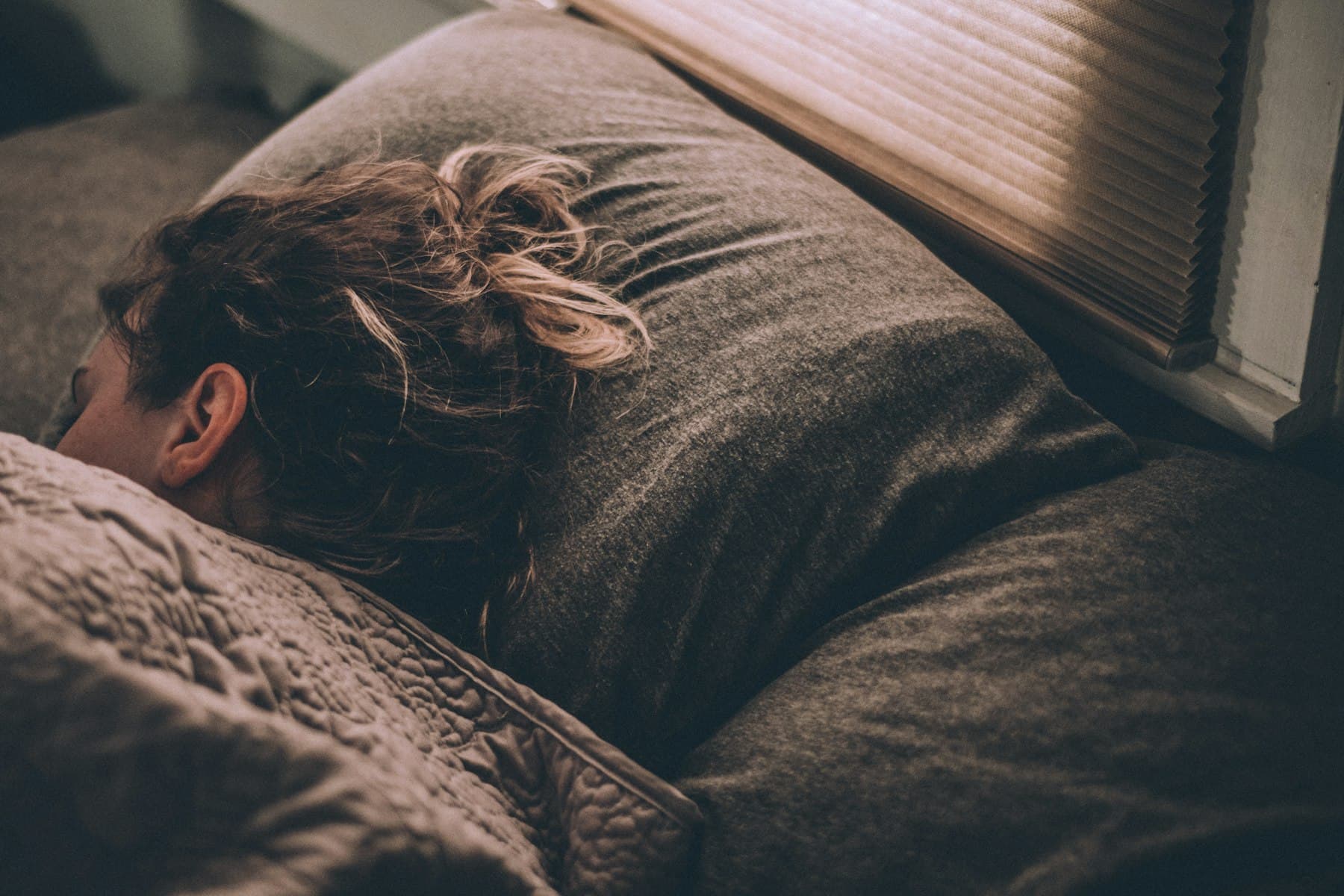 Femme endormie vue de dos, la tête sur un oreiller, dans la lumière chaude d’un store.